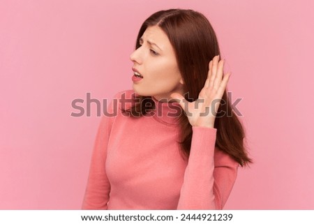Can't hear you. Attentive woman with brown hair holding hand near ear trying to listen quiet conversation, overhearing gossip, wearing rose turtleneck. Indoor studio shot isolated on pink background