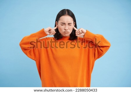 Cant listen. Fed up asian woman shuts her ears with fingers and closes eyes from loud noise, stands in orange sweatshirt over blue background.