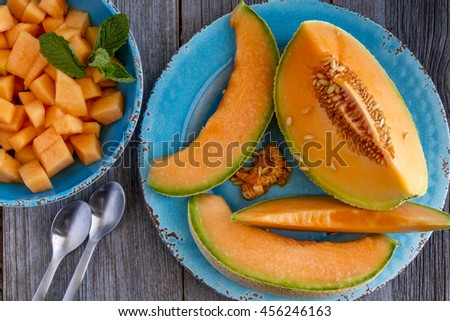 Cantaloupe melon slices sitting on blue rustic plate on wooden tabletop with two spoons and bowl filled with melon pieces