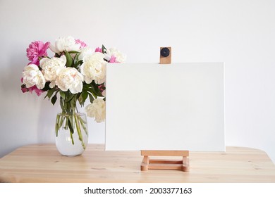 Canvas Mockup On Easel And Pink Flowers On Wooden Table On White Wall Background. Blank Artistic Canvas