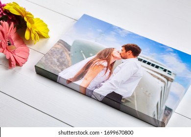 Canvas Print With Gallery Wrap Technique On White Wooden Table. Wedding Photo And Bouquet Of Gerbera Flowers. Stretched Photography, Portrait Of A Kissing Couple. Memories Of A Romantic Cruise