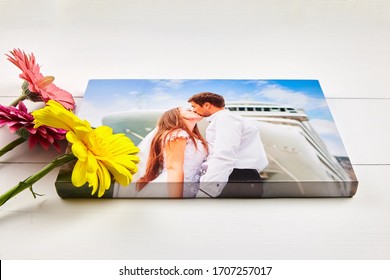 Canvas print with gallery wrap technique on white wooden table. Wedding photo and bouquet of gerbera flowers. Stretched photography, portrait of a kissing couple. Memories of a romantic cruise