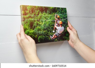 Canvas print. Photo with gallery wrap method of canvas stretching on stretcher bar. Woman hangs a color travel photography (with people on zip line) on a white wooden wall