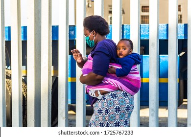 Cape Town, South Africa - 06 May 2020: African Woman Carrying Baby On Back, Wearing Face Mask During Covid 19 Corona Virus Pandemic, Trying To Survive Poverty And The Virus.