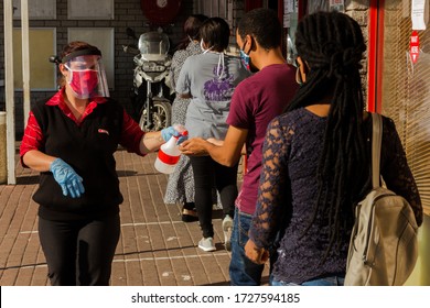 Cape Town, South Africa - May, 2020: Front Line Worker, Shop Teller Sanitizes Customers Waiting In Like With Face Shields, Corona Virus Pandemic Lock Down In Africa.