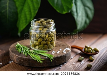 Capers in a transparent jar on a wooden stand with sea salt and rosemary. On a wooden background with green leaves.