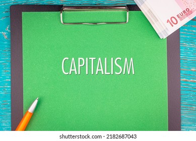 CAPITALISM - Word (text) And A Bundle Of Money, Euro Bills On A Green Background Of A Notebook, A Pen And A Wooden Table. Business Concept: Buy, Sell, Commerce (copy Space).