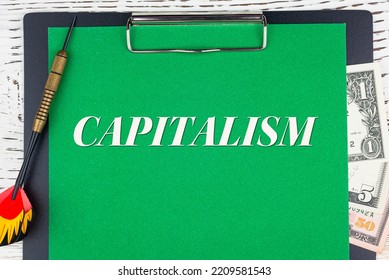 CAPITALISM - Word (text), A Dart For Darts And Dollar Bills On A Green Background Of A Notebook, A Wooden White Table. Business Concept: Buying, Selling, Commerce (copy Space).