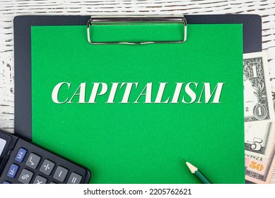 CAPITALISM - Word (text) Dollar Bills On Green Background, Pencil, Calculator And Wooden White Table. Business Concept: Buying, Selling, Commerce (copy Space).