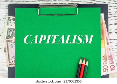 CAPITALISM - Word (text) Euro And Dollar Bills On A Green Background, Colored Pencils And A Wooden White Table. Business Concept: Buying, Selling, Commerce (copy Space).