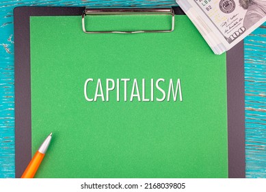 CAPITALISM - Word (text) And Money A Pack Of Dollar Bills On A Green Background Notepad, Pen. Business Concept: Buy, Sell (copy Space).