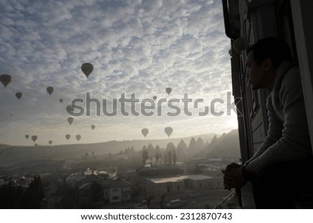 Cappadocia,Turkey- Man sits at terrace and looks up in the sky to see hot air balloons flying over the exotic rural rocky hills and fairy chimneys