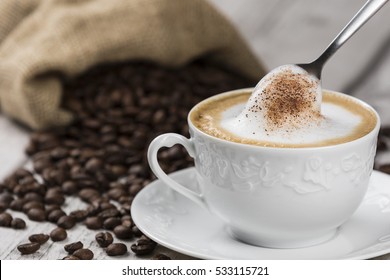 Cappuccino Coffee Cup and Spoon / Composition of Cup of Cappuccino and spoon with foam, sack of coffee beans on white wooden table
 - Powered by Shutterstock