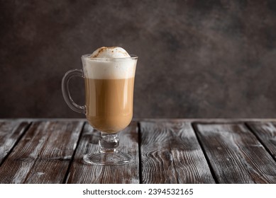 Cappuccino coffee with foam in a tall glass on a dark old wooden table. Side view, copy space.