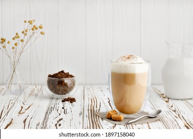 cappuccino in glass with double walls on white wooden table with coffee beans and jug of milk