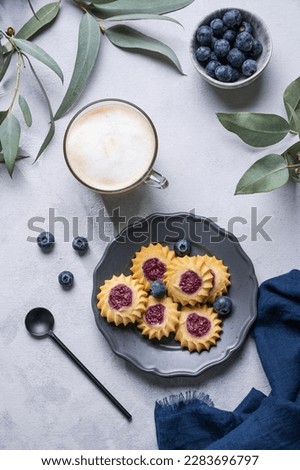 Cappuccino or latte coffee with milk foam in a cup with  homemade berry cookies in a dark plate and blueberries on a light background with eucalyptus branches. Top view.