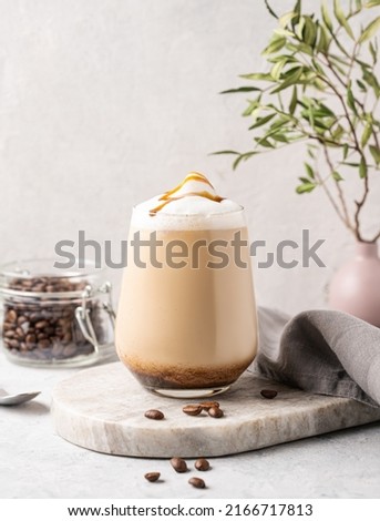 Cappuccino or latte with milk foam and caramel in a glass with coffee beans on light  marble background with branches. Front view and copy space.