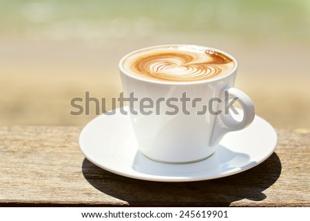 Cappuchino or latte coffe in a white cup  with heart shaped foam on wooden board with ocean on the background