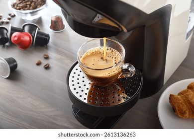Capsule coffee maker preparing espresso in a glass cup on a wooden kitchen bench with coffee capsules and pastries. Elevated view.