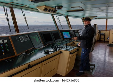 The Captain With A Beard And A Blue Cap Stands On The Bridge Of A Cargo Ship And Points To The Handheld Radio. Through The Windows You Can See The Sea And Mountains On The Horizon.