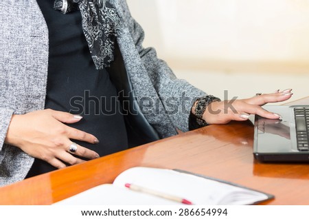 caption of pregnant womans belly wearing casual clothes at work at desk touching stomach and computer