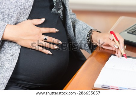 caption of pregnant womans belly wearing casual clothes at work at desk touching stomach and red pen in other hand