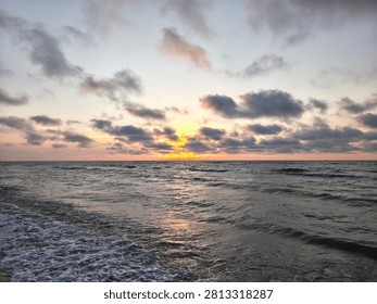 A captivating and atmospheric seascape capturing a dramatic sunset over the ocean. The horizon is illuminated by a vibrant, glowing break of bright yellow and orange light, contrasting sharply with the moody, textured dark grey clouds scattered across the pale evening sky. Below, the dark, churning waters of the sea reflect the warm hues of the setting sun, creating a subtle, shimmering pathway of light on the water's surface. In the foreground, dynamic waves roll toward the viewer, breaking into frothy white sea foam as they wash onto the shoreline. This serene yet powerful coastal landscape perfectly encapsulates the tranquil beauty of twilight and the raw energy of the ocean. It is an ideal background image for themes related to nature, travel, meditation, coastal environments, and dramatic weather patterns.