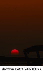 A captivating image of a deep red sun setting over the ocean horizon, with a dramatic gradient sky and a dark silhouette of a wooden structure in the foreground, evoking peace and wonder.