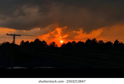 A captivating image of a dramatic sunset with a fiery orange sky, partially obscured by dark, looming clouds. The sun is setting behind a silhouette of trees, with power lines and a utility pole. - Powered by Shutterstock