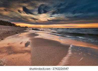 A captivating long-exposure photograph capturing the dramatic beauty of a sunset over a tranquil sandy beach. The sky is a breathtaking canvas of deep blues and vibrant oranges, accented by swirling, painterly clouds. The low sun casts a golden glow across the horizon, which is beautifully reflected in the wet, smooth sand in the foreground, where gentle, frothy waves recede, creating soft, ethereal patterns. A distant line of trees and scattered rocks along the shore complete this peaceful coastal landscape.