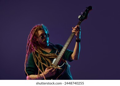 Captivating sight of man with dreadlocks, musician expressing his emotions through music, guitar playing against dark purple background in neon light. Concept of music, performance, festival, concert - Powered by Shutterstock