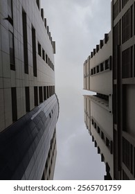 A captivating upward view between two tall modern buildings, showcasing symmetry, clean geometric lines, and depth. The cloudy sky adds a subtle yet dramatic contrast, enhancing the urban aesthetic.