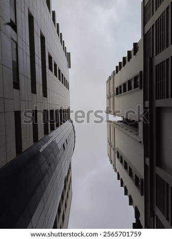 A captivating upward view between two tall modern buildings, showcasing symmetry, clean geometric lines, and depth. The cloudy sky adds a subtle yet dramatic contrast, enhancing the urban aesthetic.