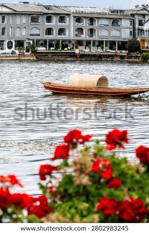 A captivating vertical waterscape looking across the calm, shimmering expanse of Erhai Lake in Dali, Yunnan, China. Vibrant red geranium blossoms frame the lower foreground in a gentle blur, adding artistic depth to the composition. In the center, a solitary traditional wooden boat with a rustic woven bamboo shelter floats quietly on the water. The background reveals the stylish, arched architecture of coastal guesthouses and boutique resorts, capturing the elegant, leisurely lifestyle of southwestern China's famous vacation retreats.