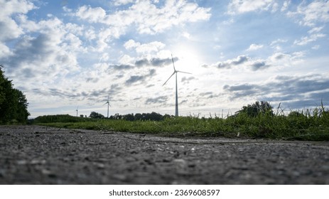 A captivating very low angle view captures an asphalt road flanked by lush grass, leading to a majestic electric windmill standing tall. The setting sun creates a stunning silhouette. Electric - Powered by Shutterstock