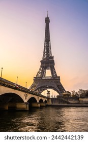 A captivating view of the Eiffel Tower and a bridge over the Seine River at sunrise. The sky is painted with warm hues, casting a serene atmosphere over Paris, France.