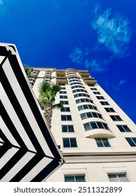 A captivating view from a poolside, looking up at a tall hotel that towers against a clear blue sky. The scene is framed by elegant palm trees and a stylish black and white striped umbrella.