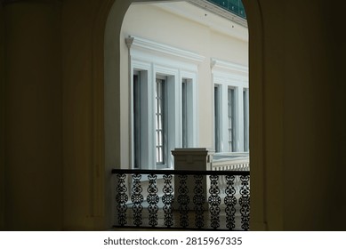 A captivating view through a shadowed archway framing ornate black metal railing and classically designed windows. Architectural details like the elegant molding and patterned balustrade evoke a sense of timeless charm. The interplay of light and shadow enhances this sophisticated building perspective.