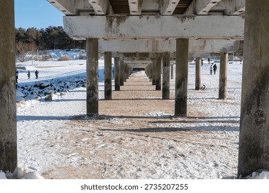 A captivating view from underneath a robust concrete pier, showcasing a rhythmic repetition of massive support pillars extending into the horizon. The ground is a serene mix of sand and pristine snow, illuminated by the bright winter sun casting dramatic, elongated shadows. In the background, people enjoy a leisurely walk across the vast, snow-covered coastal landscape, framed by a dense line of evergreen trees and a clear, azure sky.