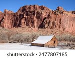 Captivating winter scene of the historic Fruita Barn at Capitol Reef National Park, Utah. Snow-covered ground, red rock cliffs, and clear blue sky create a picturesque and serene landscape - USA