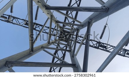A captivating worm's-eye view looking directly up through the center of a high-voltage electrical transmission tower. The image highlights the complex, symmetrical geometric patterns of the galvanized steel lattice structure converging towards the top. Ceramic power insulators and heavy-duty power lines are visible against a clear blue sky background. This striking industrial infrastructure photography perfectly illustrates concepts of electricity distribution, energy networks, engineering, and modern power grids.