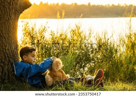 Capture the essence of childhood innocence and wonder in this heartwarming photo. A 10-year-old boy sits against a tree by the serene forest lake, his cherished cuddly bear by his side. As the sunset