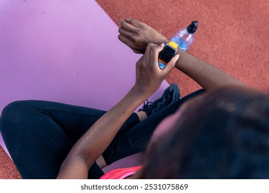 Captured from above, the African American woman sits on her exercise mat, her focus on her hands and smartwatch.  - Powered by Shutterstock