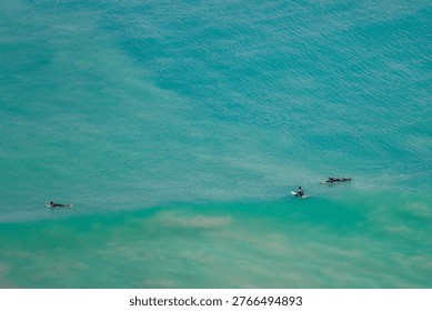 Captured from an elevated perspective, this image showcases the serene beauty of the ocean in Hawaii, United States. The turquoise water stretches out in a vast expanse, its surface gently rippling with subtle textures. Three individuals on surfboards are sparsely scattered across the scene, each a small silhouette against the immensity of the sea. One surfer is positioned further out in the open water, while the other two are closer to a zone where the water appears shallower and lighter in color, hinting at a reef or sandbar beneath. The lighting suggests a bright, sunny day, accentuating the vibrant hues of the water and creating a sense of tranquility and vastness. The composition emphasizes the relationship between humanity and nature, with the surfers appearing small and solitary against the backdrop of the endless ocean. The overall mood is one of peace and solitude, capturing the essence of a tropical paradise and the spirit of surfing.