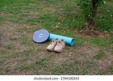 Captured from a high right-angled perspective, this view highlights a blue rolled fitness mat, a leaning kinetic twister, and brown cream sneakers on garden turf.