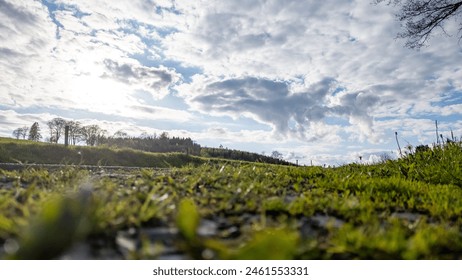 Captured from a low angle, this image features the lush, grassy terrain of the Hautes Fagnes region under a dynamic sky filled with scattered clouds. The foreground shows a close-up of greenery