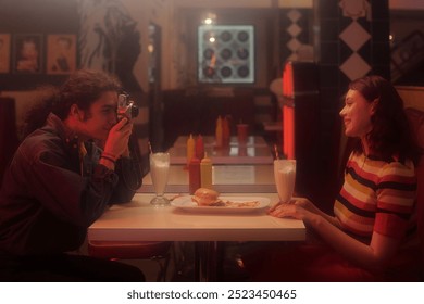 Capturing candid photograph in retro-themed diner setting with friends. One person holding vintage camera, another person enjoying milkshake and burger - Powered by Shutterstock