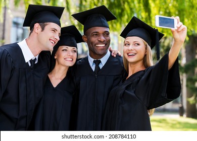 Capturing A Happy Moment. Four College Graduates In Graduation Gowns Standing Close To Each Other And Making Selfie