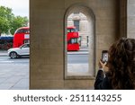 Capturing London’s Iconic red buses: A tourist’s snapshot through historic arches on a bustling city street in the UK