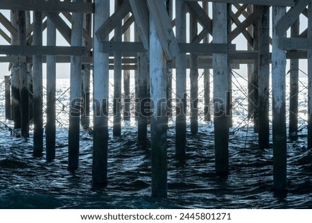 Capturing the intricate patterns of light and shadow, this image showcases the robust wooden structure of a pier with its pillars and crossbeams plunging into the dark, swirling waters below, offering